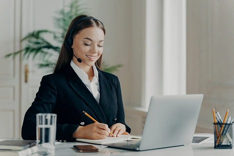 Aydınlık bir ofiste, masasında dizüstü bilgisayarıyla çalışırken notlar alan genç bir profesyonel kadın.